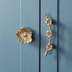 May include: A gold-toned metal cabinet knob shaped like a single flower and a matching vertical cabinet pull handle featuring three small flower motifs along a curved branch, both mounted on a solid light blue paneled cabinet door surface.