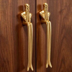 May include: Two polished gold-toned metal cabinet handles shaped like stylized figures sitting with their hands near their faces. The figures have elongated legs extending downward, mounted vertically on a dark brown wood grain surface.