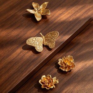 May include: A collection of four gold-toned metal decorative pins featuring a butterfly and three distinct floral designs. Each piece displays intricate embossed textures and detailed petal or wing patterns with a polished metallic finish.