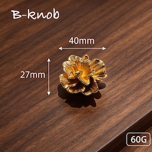 May include: A gold-colored B-knob shaped like a blooming flower with textured petals. The knob measures 40mm in width and 27mm in height, with a weight of 60g. The design features layered, organic petal shapes with a metallic finish.