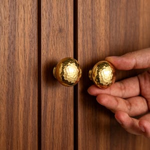 Op de afbeelding: Twee ronde, goudkleurige kastknoppen met een gehamerde, gefacetteerde textuur. De knoppen zijn gemonteerd op een mediumbruin houten oppervlak met verticale nerf en hebben een reflecterende metalen afwerking die het licht opvangt.