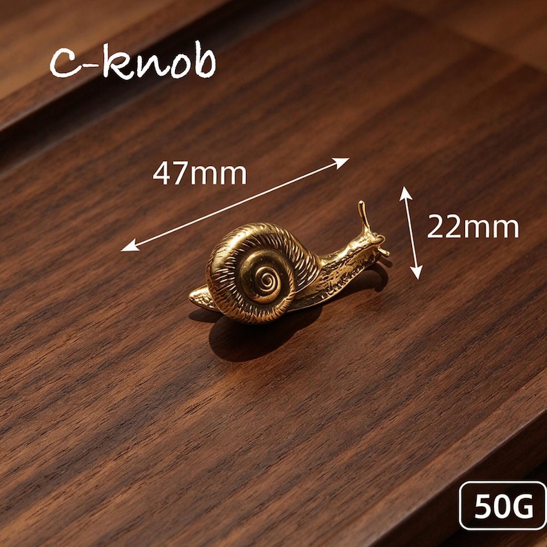 May include: A gold-toned metal snail-shaped knob labeled c-knob. The snail measures 47mm in length and 22mm in height, with a weight of 50g. The design features a detailed spiral shell and two antennae, rendered in a metallic finish.
