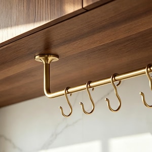 May include: A brass-toned metal rail mounted to the underside of a wooden cabinet, featuring a series of removable S-shaped hooks. The hardware includes a circular mounting plate and a horizontal rod designed for hanging various kitchen items.