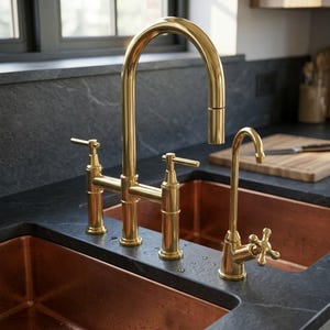 May include: A polished gold kitchen tap with a high-arc spout and two lever handles, paired with a separate smaller gold filtered water tap featuring a cross-handle design, all mounted on a dark stone worktop above a copper sink.