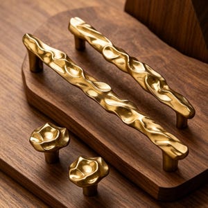 May include: Two elongated gold-toned cabinet pulls with a wavy, textured surface and two matching round cabinet knobs with a similar irregular, organic, and rippled finish. The hardware pieces feature a polished metallic sheen throughout.