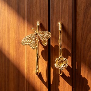 May include: Two gold-colored metal cabinet handles mounted on a dark wood door. The left handle features a detailed butterfly design, while the right handle features a floral design. Both handles have a slender, organic, branch-like vertical bar.