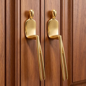 May include: Two gold-coloured metal cabinet pulls, each shaped like a stylised human figure sitting with long, slender legs extending downwards. The figures have smooth, rounded heads and torsos, mounted vertically on a wooden cabinet door surface.