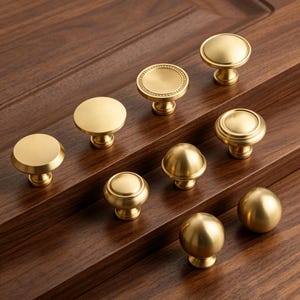 May include: A collection of eight round cabinet knobs in a brushed gold finish. The knobs feature various designs including flat circular tops, domed shapes, and tiered edges, all mounted on small cylindrical bases with a metallic sheen.