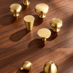 May include: A collection of gold-colored metal cabinet knobs in various shapes, including round, mushroom, and tapered designs. The knobs feature a smooth, brushed finish and are displayed in a group, with two additional spherical knobs shown below.