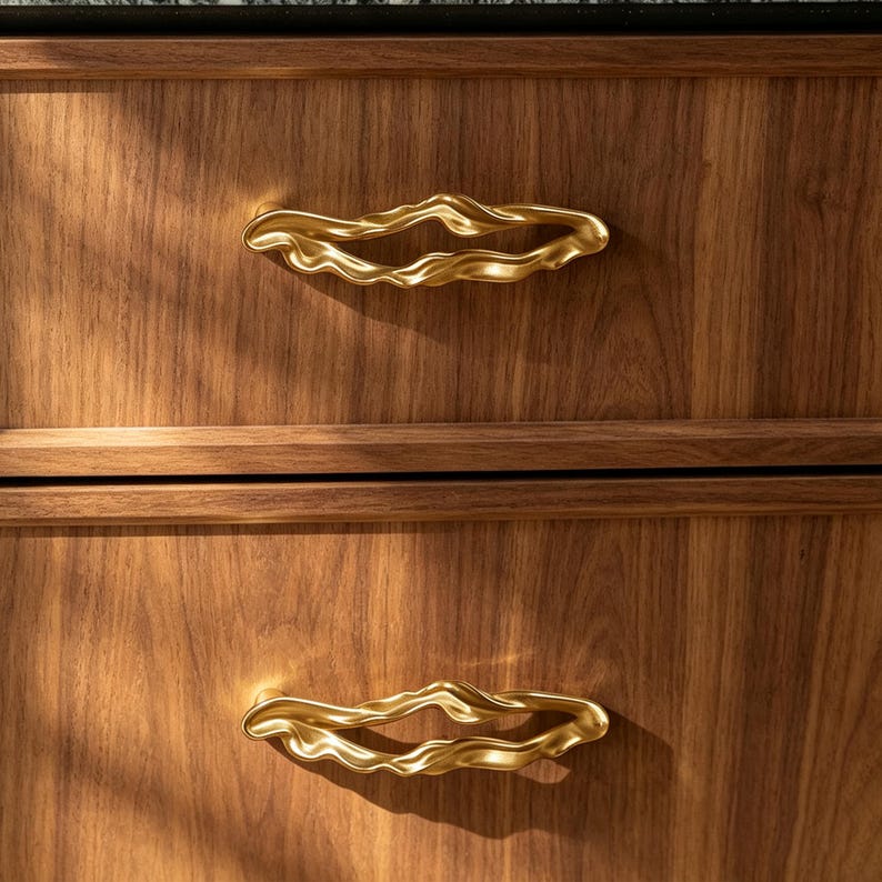 Puede incluir: Dos tiradores de caj&oacute;n de metal dorado con una forma org&aacute;nica, irregular y ondulada, fijados a un armario de madera con un patr&oacute;n de veta visible. Los tiradores presentan una superficie lisa y reflectante con un centro hueco, montados horizontalmente.