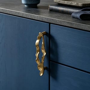 Puede incluir: Un tirador de armario de metal dorado con una forma org&aacute;nica, fluida e irregular. El tirador est&aacute; montado verticalmente en una puerta de armario de madera azul oscuro, con una superficie lisa y reflectante que contrasta con el acabado azul mate.