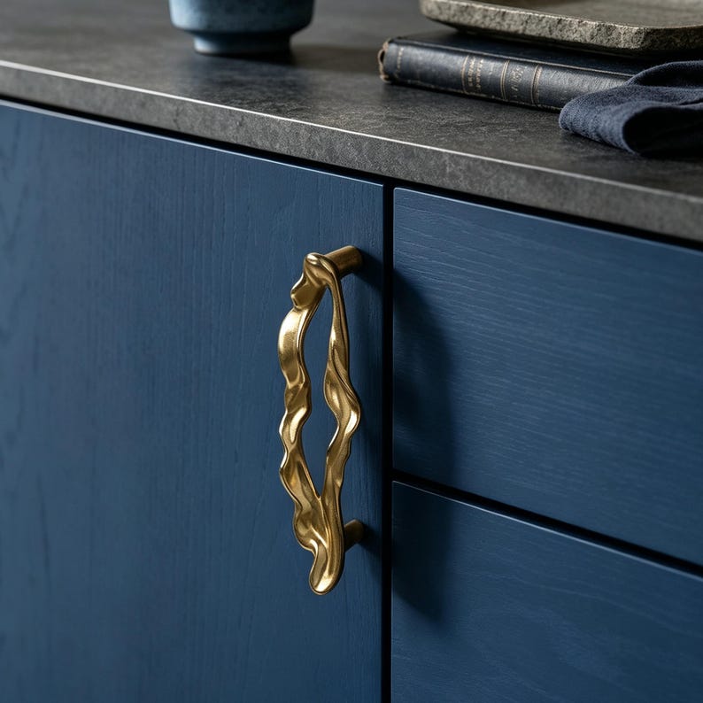 Puede incluir: Un tirador de armario de metal dorado con una forma org&aacute;nica, fluida e irregular. El tirador est&aacute; montado verticalmente en una puerta de armario de madera azul oscuro, con una superficie lisa y reflectante que contrasta con el acabado azul mate.