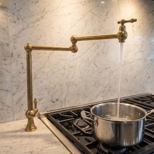 Könnte beinhalten: Ein an der Wand montierter Topffüller-Wasserhahn aus Messing mit einem beweglichen, mehrgelenkigen Arm, der über einen Edelstahlherd ragt. Der Wasserhahn hat ein Vintage-Finish und gibt einen stetigen Wasserstrahl in einen Metalltopf ab.