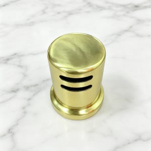 以下が含まれることがあります： 平らな上面と底面を持つ、真鍮色の円筒形の物体。円筒の側面には2つの水平なスリットが切り込まれています。この物体は、白と灰色のマーブル模様の表面に置かれています。