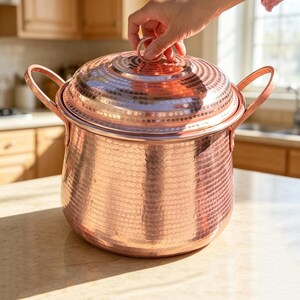 Puede incluir: Una olla grande y redonda de cobre martillado con tapa a juego y dos asas laterales. La superficie metálica presenta una textura uniforme de hoyuelos en todo el exterior de la olla y la tapa, reflejando la luz sobre su acabado metálico.