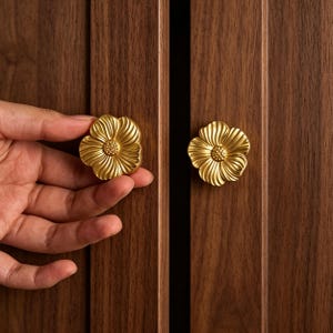 May include: Two gold-colored metal cabinet knobs shaped like five-petaled flowers with textured centers and radiating petal lines. The knobs are mounted on dark brown wooden cabinet doors with vertical grain patterns and a central seam.