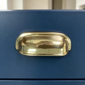 May include: A polished brass cup pull handle mounted on a matte navy blue drawer front. The hardware features a smooth, curved, semi-circular design with a reflective metallic finish and two visible screw attachment points on the sides.