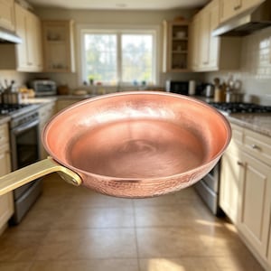 May include: A round, hammered copper skillet with a polished interior surface and a long, straight brass handle attached by two rivets. The exterior of the pan features a textured, dimpled finish that catches the light, contrasting with the smooth, metallic handle.