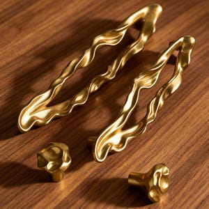 May include: Two elongated, wavy gold cabinet pulls with a textured, organic finish and two matching small, round, irregular gold cabinet knobs. The hardware features a matte metallic surface with soft curves and a fluid, molten-like appearance.