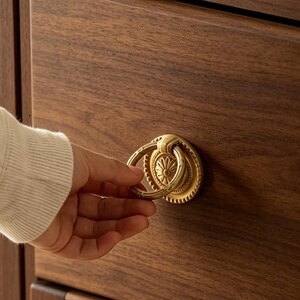 May include: A circular brass ring pull handle mounted on a dark brown wooden drawer. The handle features a decorative backplate with a radial floral pattern and a smooth, polished finish, contrasting against the natural wood grain texture.