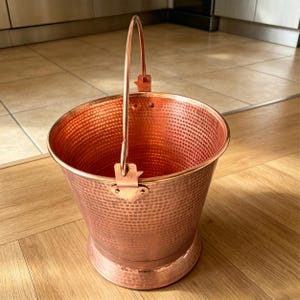 May include: A polished copper bucket featuring a distinct hammered texture across its exterior surface. The vessel includes a sturdy, upright metal handle attached with riveted brackets, a wide circular opening, and a reinforced base rim.