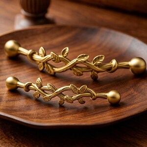 May include: Two gold-toned metal cabinet pulls featuring a decorative vine and leaf design. Each handle has a smooth, rounded ball at both ends and a central section shaped like intertwined branches with small, detailed leaves along the length.