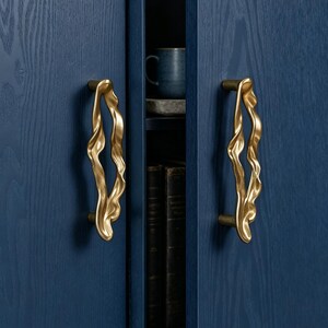 Puede incluir: Dos tiradores de armario verticales de lat&oacute;n con una silueta org&aacute;nica, fluida y ondulada, montados en puertas de armario de madera azul oscuro. Los tiradores presentan un acabado met&aacute;lico pulido con curvas irregulares y onduladas.