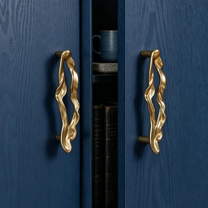 Puede incluir: Dos tiradores de armario verticales de lat&oacute;n con una silueta org&aacute;nica, fluida y ondulada, montados en puertas de armario de madera azul oscuro. Los tiradores presentan un acabado met&aacute;lico pulido con curvas irregulares y onduladas.