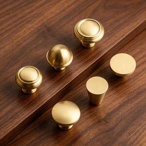 May include: A collection of six brass cabinet knobs in two distinct shapes. Three knobs feature a rounded, tiered profile with a smooth finish, while the other three are flat-topped, cylindrical knobs with a brushed gold-toned metallic surface.