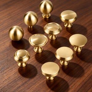May include: A collection of nine brass cabinet knobs in various shapes, including spherical, flat-topped, and tiered designs. Each knob features a smooth, brushed metallic finish with a circular base and a central stem for mounting to furniture.