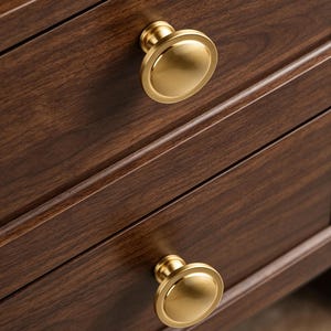 May include: Two circular cabinet knobs with a brushed brass finish attached to a dark brown wooden drawer front. The knobs feature a smooth, slightly domed center with a flat, wide rim, providing a clean and functional hardware design.
