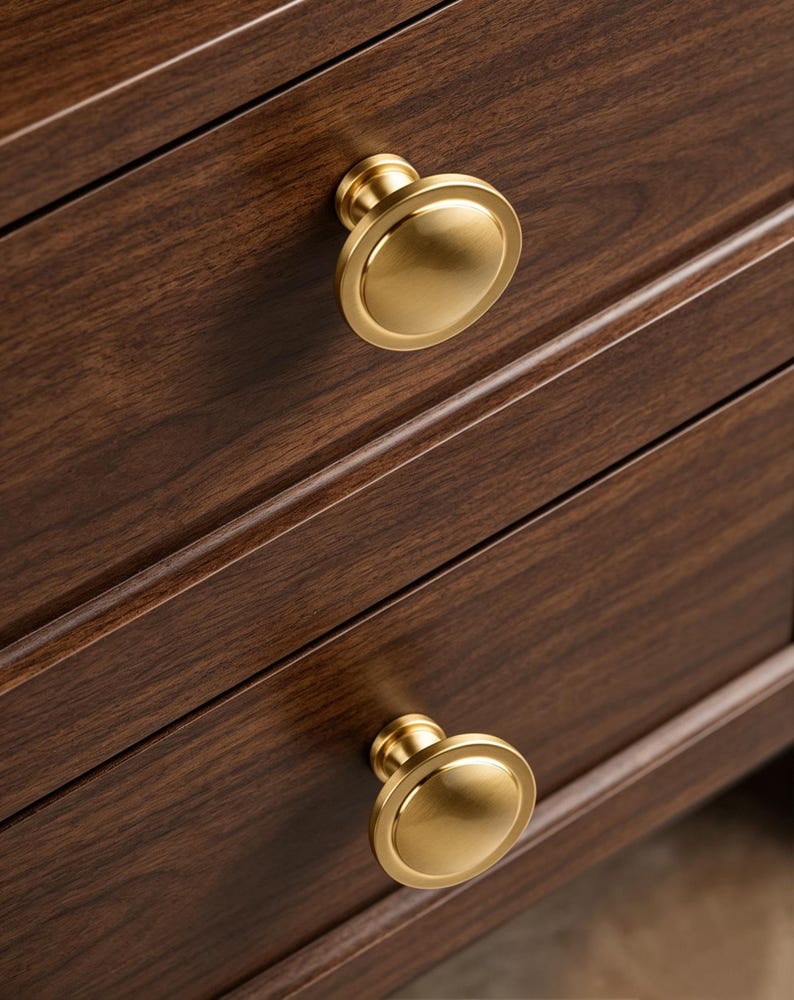 May include: Two circular cabinet knobs with a brushed brass finish attached to a dark brown wooden drawer front. The knobs feature a smooth, slightly domed center with a flat, wide rim, providing a clean and functional hardware design.