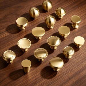 May include: A collection of fifteen brass-toned metal cabinet knobs in various circular and spherical shapes. Each knob features a smooth, brushed finish with a consistent golden hue, showcasing different profiles including flat tops, rounded domes, and tapered bases.