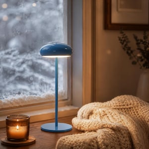 May include: A blue table lamp with a round base and a mushroom-shaped top. The lamp is on a wooden surface near a window with a snowy view. A lit candle in a glass jar and a knitted blanket are also visible.