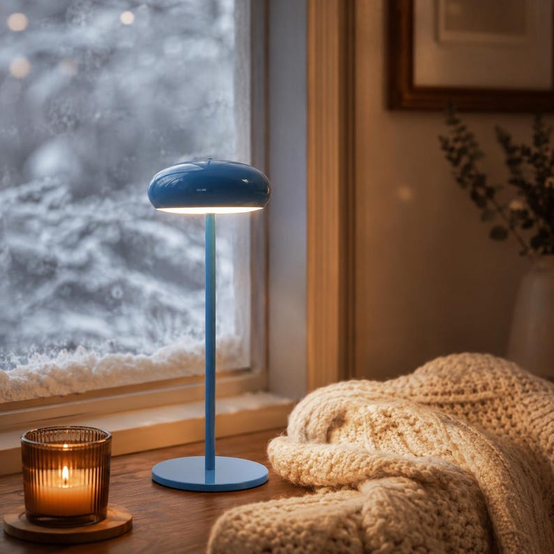 May include: A blue table lamp with a round base and a mushroom-shaped top. The lamp is on a wooden surface near a window with a snowy view. A lit candle in a glass jar and a knitted blanket are also visible.