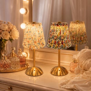 May include: Two decorative table lamps with pleated floral shades. The lamps have a gold-colored base and stem. The shades feature a colorful floral pattern with red, blue, and yellow flowers. The lamps are on a white surface.