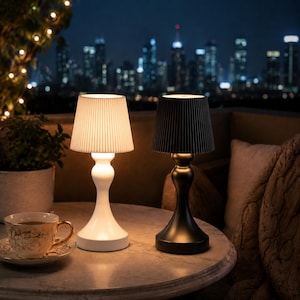 Pode incluir: Duas luminárias de mesa sem fio com abajures plissados ​​em uma mesa redonda. Uma luminária é branca, a outra preta. Uma xícara e um pires estão sobre a mesa. O fundo apresenta um horizonte noturno com luzes de corda.