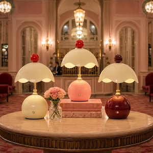May include: Three table lamps with scalloped cream-coloured shades and spherical bases in cream, pink, and dark red. Each lamp features a small, round finial on top of the shade and a gold-toned stem. A small glass vase with pink roses sits in the centre.