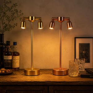 May include: Two metal table lamps with a central vertical stem and two downward-facing light heads. One lamp has a brushed gold finish, and the other has a copper finish. Both lamps feature a circular base and emit a warm, soft glow.