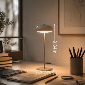 May include: A modern, minimalist table lamp with a mushroom-shaped shade. The lamp is 9.84 inches tall and has a base diameter of 3.93 inches. It is a neutral color and is turned on, illuminating the surrounding area. The lamp is on a desk with office supplies.