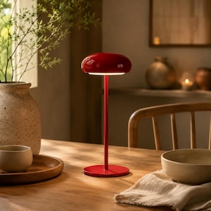 May include: A red table lamp with a rounded top and a slender stem. The lamp is made of metal and has a circular base. The lamp is turned on, emitting a soft, warm light. The lamp is on a wooden table.