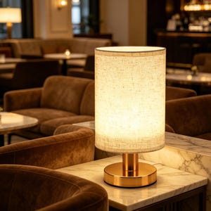 May include: A cylindrical table lamp with a gold base and a beige fabric shade. The lamp is lit, casting a warm glow. The lamp is on a marble-topped table in a lounge setting with brown velvet seating.