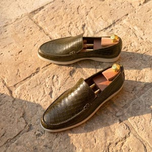 Peut inclure: Deux paires de mocassins en cuir vert olive avec une texture peau de crocodile. Les chaussures ont un design à enfiler avec une semelle blanche et des embauchoirs en bois à l'intérieur. Les mocassins sont exposés sur une surface en pierre.
