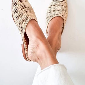 May include: A pair of beige woven mules featuring a pointed toe and a flat brown sole. The upper part of the footwear is constructed from a textured, open-weave material in a light tan color, providing a breathable design with a slip-on style.