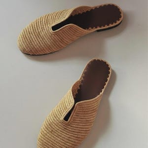 Könnte beinhalten: Ein Paar beigefarbene, geflochtene Slipper mit geschlossener, abgerundeter Zehenpartie, einem tiefen V-Ausschnitt am Rist und einer dunkelbraunen Innensohle mit gewelltem Rand. Das Obermaterial besteht aus einer strukturierten Strohoptik.