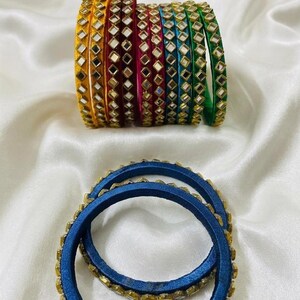 May include: A collection of circular bangles featuring a variety of vibrant colors including yellow, red, pink, blue, and green. Each bangle is wrapped in thread and decorated with a series of small, square-shaped reflective mirror accents.
