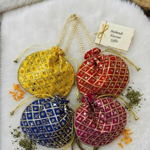 May include: Four colorful, embellished drawstring pouches in yellow, red, blue, and magenta. Each pouch features a geometric pattern with gold accents and small mirrors. The pouches have pearl-beaded handles and are arranged on a white, fluffy surface.