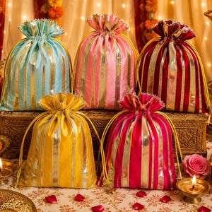 May include: Five drawstring pouches in light blue, pink, deep red, yellow, and magenta. Each pouch features vertical metallic gold or silver stripes, a gathered top with a drawstring closure, and a shimmering fabric finish.