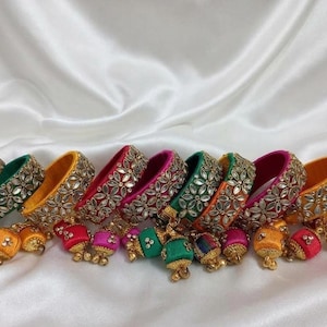 May include: A collection of circular bangles wrapped in vibrant silk thread in shades of yellow, green, pink, and orange. Each bangle is adorned with intricate floral-shaped metallic embellishments and features small dangling charms.