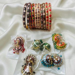 May include: A set of ten metal bangles featuring colorful floral enamel patterns in shades of navy, red, green, and cream, alongside five pairs of bell-shaped jhumka earrings in pink, green, yellow, white, and blue, each enclosed in clear plastic.